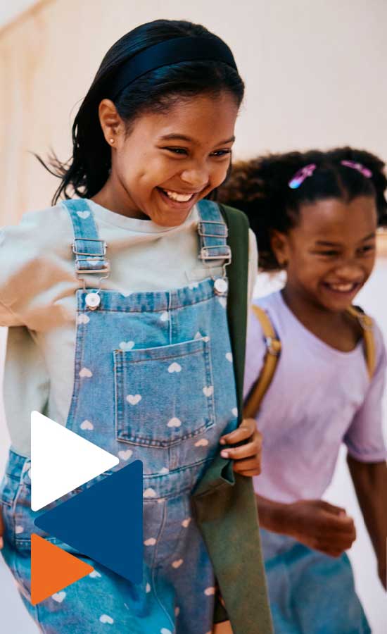 Two young girls in school clothes are smiling and walking. The girl in the foreground is looking over her shoulder at the girl behind her.