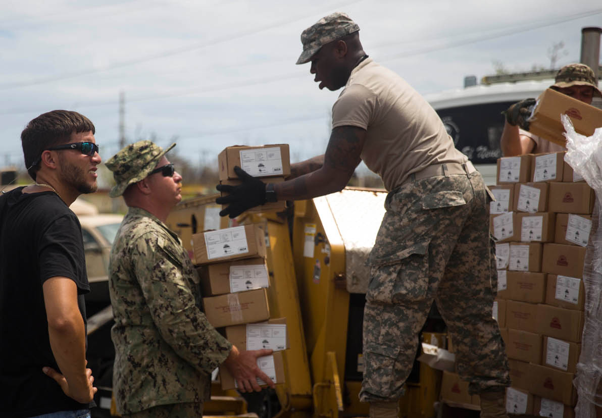 Image of two warfighters loading a truck full of small boxes with one civilian onlooker