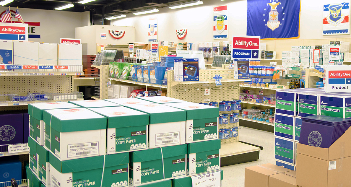 Walls and racks of office supplies inside a base supply center with AbilityOne logos throughout. 